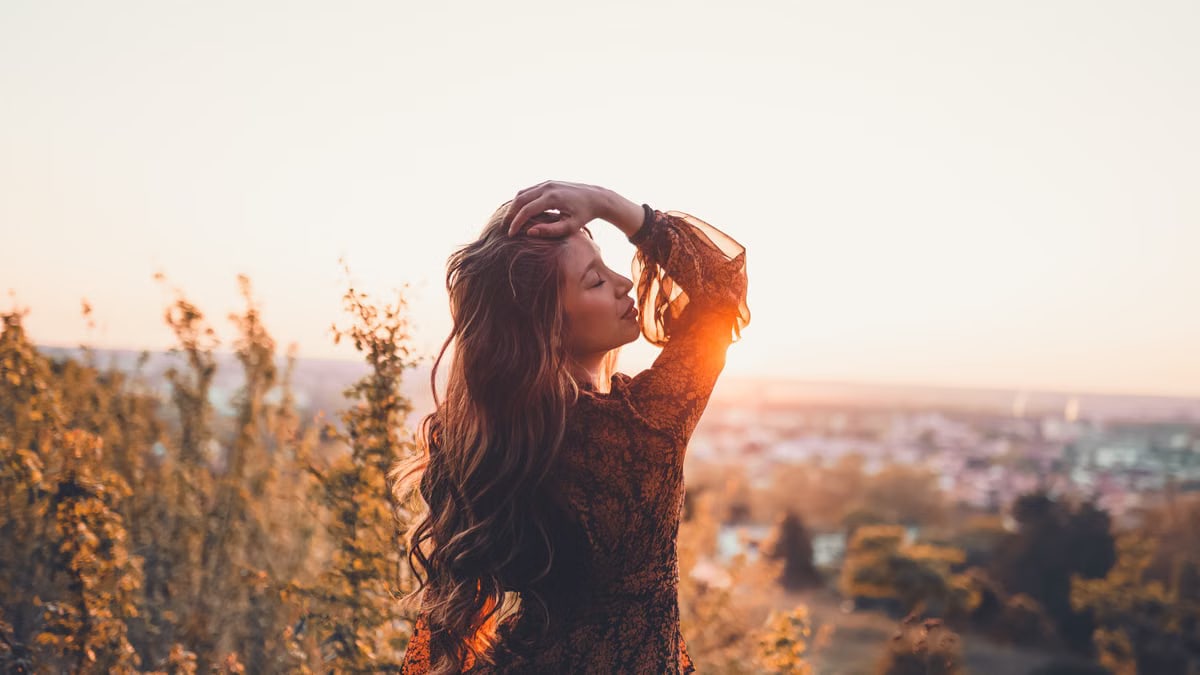 Back view of young serene female with closed eyes and wavy hair standing in grass at sunset